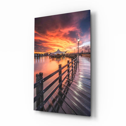 Angled view of glass wall art depicting a wooden pier railing extending toward a dramatic orange and purple sunset over water and distant cityscape.