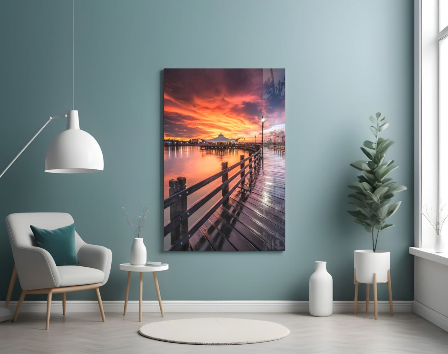 Glass wall art displaying a wooden pier at sunset with orange and pink sky reflected in calm water, mounted on teal wall above chair.
