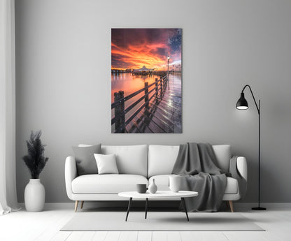 Bridge of Love Glass Wall Art displayed above white sofa showing wooden pier at dramatic orange sunset over water.