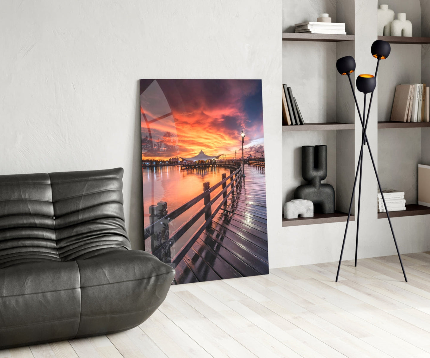 Glass wall art displaying a wooden pier at sunset with orange and pink sky reflected in water, displayed in modern living room setting.