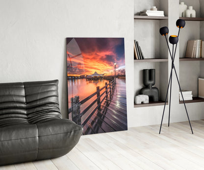 Glass wall art displaying a wooden pier at sunset with orange and pink sky reflected in water, displayed in modern living room setting.