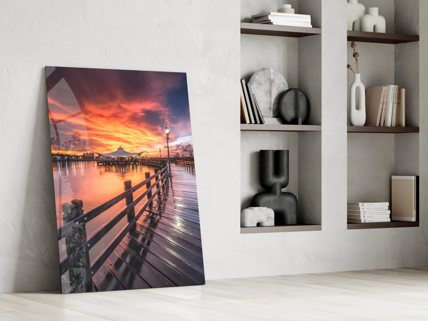 Glass wall art leaning against white wall displays vibrant sunset over wooden pier with orange sky and water reflections.