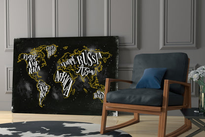 Black canvas world map with gold outlines and white text labels displayed in modern living room next to wooden chair.