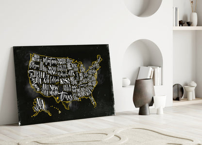 Black glass USA map with gold state names and border leaning against white wall beside decorative objects.