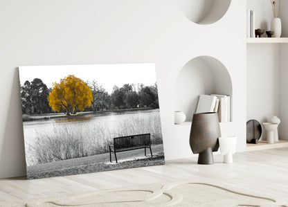Glass wall art leaning against white wall displays black and white landscape with single yellow tree and bench beside water.