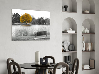 Yellow tree with golden foliage beside water and bench, displayed as glass wall art in minimalist interior setting.