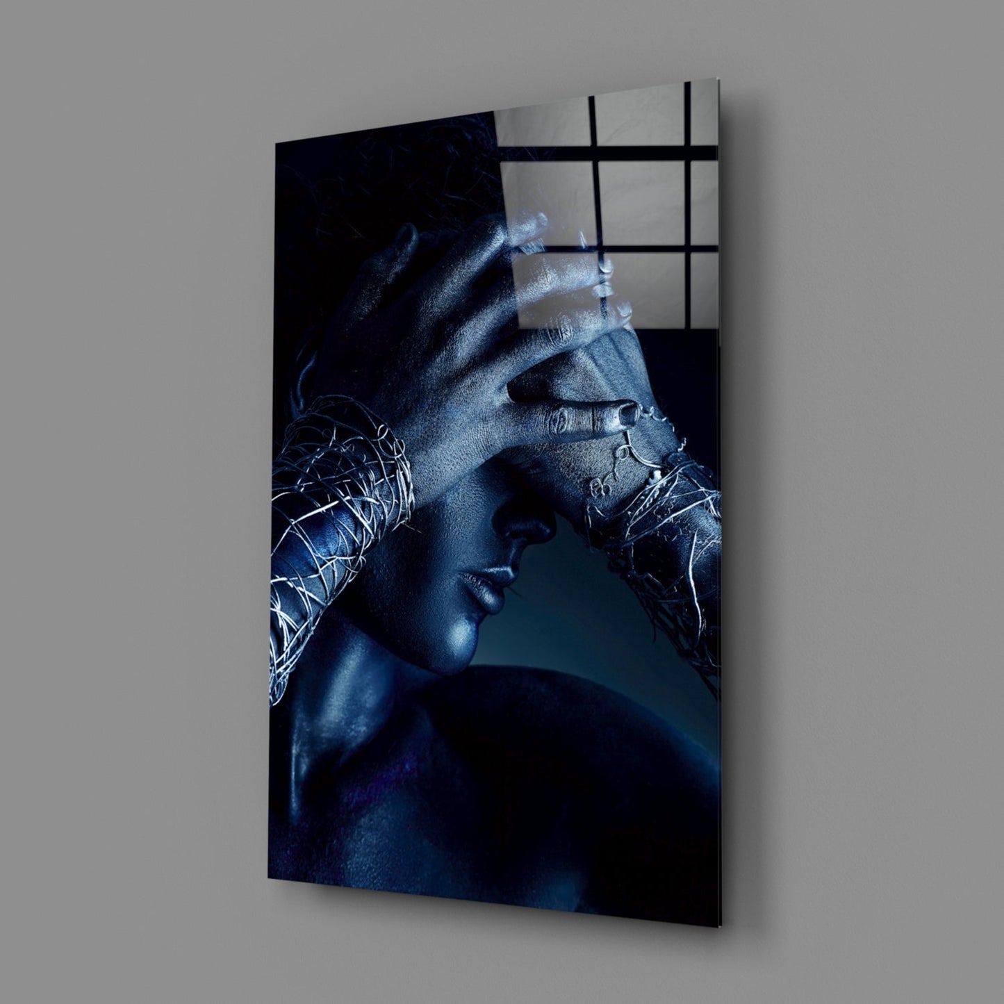 Angled view of Deep Thought Glass Wall Art showing a blue-toned figure with hands near face, metal grid overlay, mounted on gray wall.