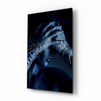 Profile view of a person with blue-toned skin and hands covering their face, displayed on glass wall art against dark background.