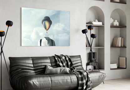 Surreal glass art depicting a figure with a colorful hot air balloon instead of a head against a cloudy sky, displayed above a black leather sofa in a modern living room.