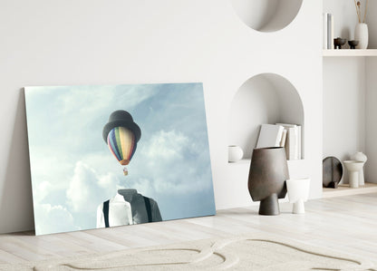 Glass wall art leaning against white wall showing man with bowler hat gazing at colorful hot air balloon in cloudy sky.