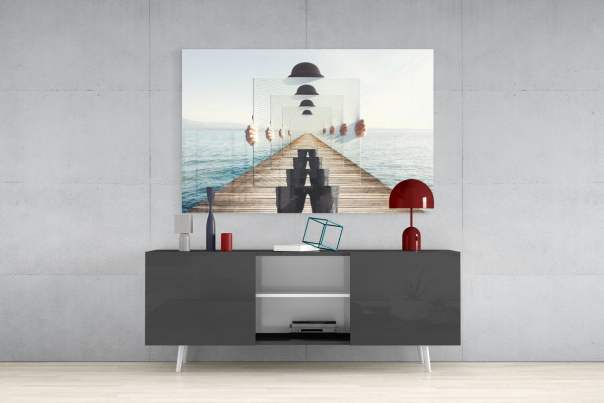 Surreal glass wall art depicting a recursive pier with bowler hats displayed above a dark media console with red lamps and decorative objects.