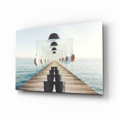 Angled product shot of glass wall art depicting a wooden pier extending toward calm water with recursive layers of translucent panels and black bowler hats diminishing into distance.
