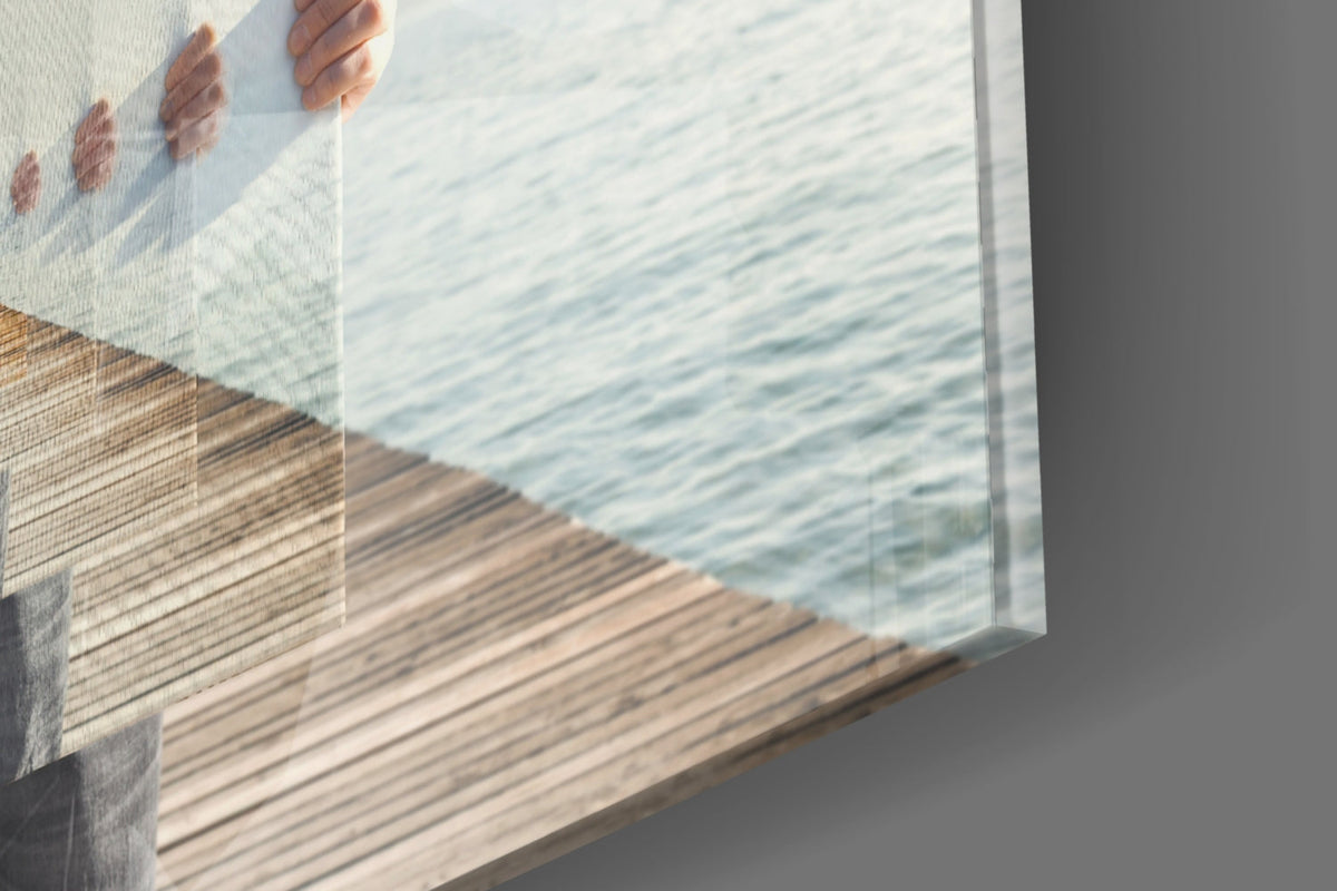 Angled close-up of glass wall art displaying a wooden pier and water scene with hand holding top edge against gray wall.