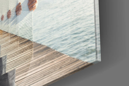 Angled close-up of glass wall art displaying a wooden pier and water scene with hand holding top edge against gray wall.