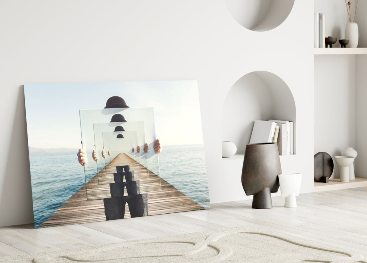Surreal glass wall art leaning against white wall in minimalist room, showing wooden pier with recursive bowler hats and figure, ocean background.