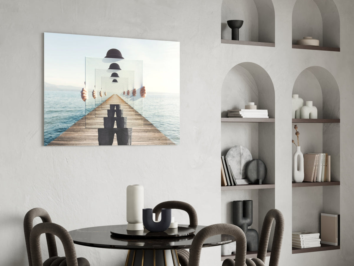Surreal glass wall art depicting a figure in dark clothing on a wooden pier with receding mirrors showing repeated bowler hats, displayed above a black round table in a minimalist interior.
