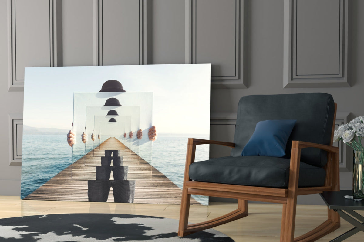 Surreal glass wall art depicting a figure in bowler hat on wooden pier with recursive mirror reflections and ocean backdrop, displayed in modern room with wooden chair.