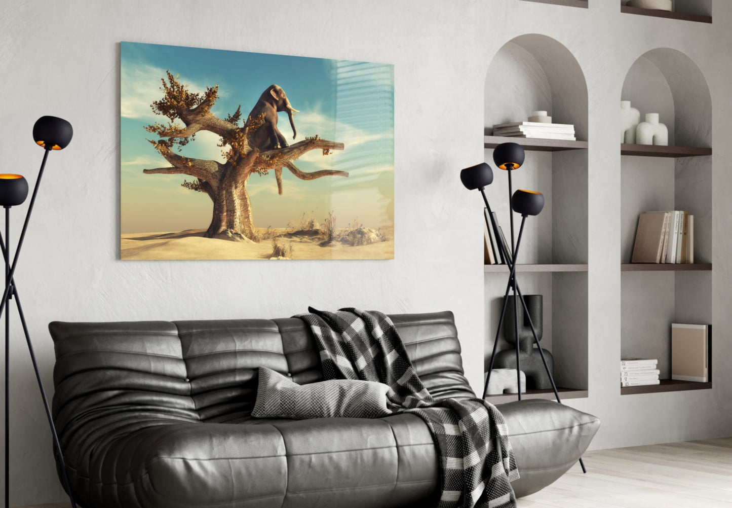 Glass wall art depicting an elephant balancing on a twisted tree branch in a desert landscape, displayed above a black leather sofa in a modern living room.