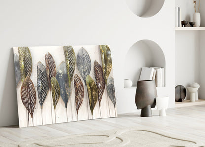 Glass wall art featuring metallic feathers in gold, brown, and gray tones displayed against white interior wall with shelving and decorative objects.