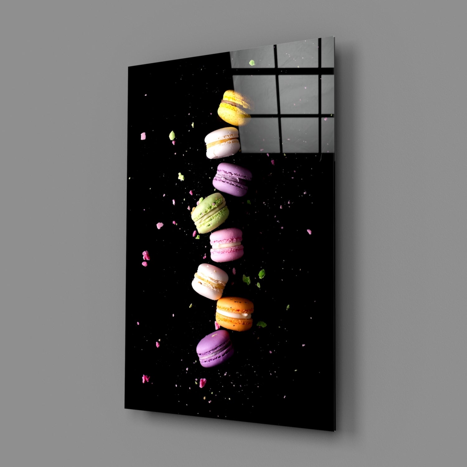 Angled view of black glass wall art panel displaying colorful macarons arranged vertically with scattered crumbs against dark background, mounted on gray wall.