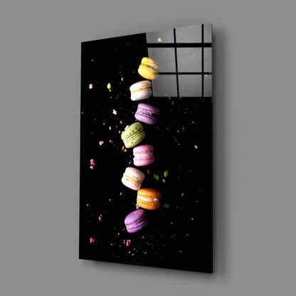 Angled view of black glass wall art panel displaying colorful macarons arranged vertically with scattered crumbs against dark background, mounted on gray wall.