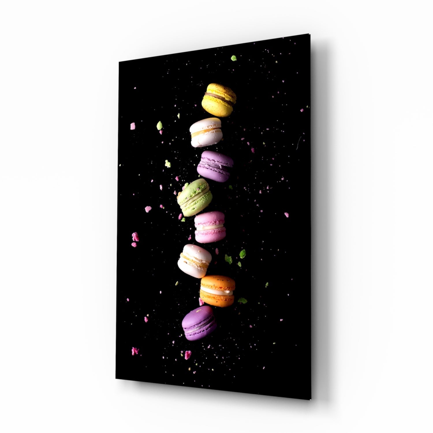 Angled view of glass wall art displaying a vertical stack of colorful macarons against a black background with scattered crumbs.