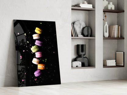 Colorful macarons stacked vertically on black glass art leaning against white wall beside wooden shelves.