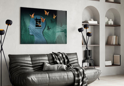 Glass wall art displaying a teal textured head silhouette with orange butterflies and a window, hung above a black leather sofa in a modern living room.