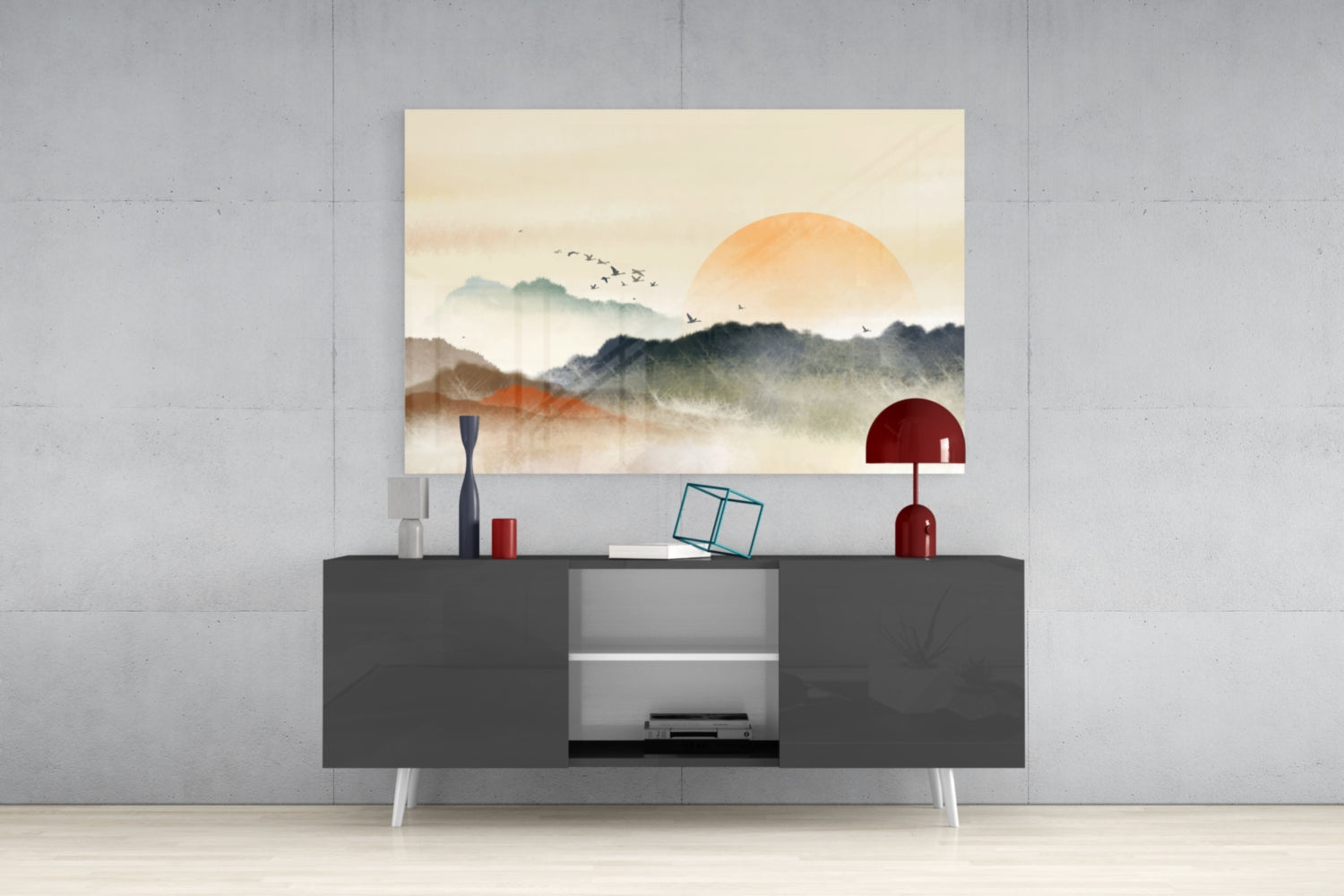 Abstract landscape glass wall art depicting misty mountains and large orange sun displayed above dark gray media console with decorative accessories.