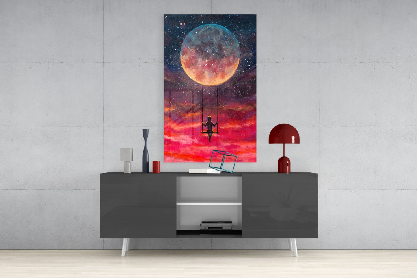 Freedom Glass Wall Art depicting a figure on a swing beneath a luminous moon against a red and orange sky, displayed above a dark console table in a modern interior setting.