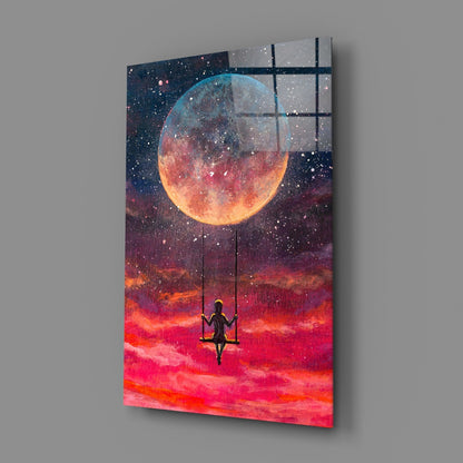 Angled 3/4 view of glass wall art depicting a silhouetted figure on a swing beneath a glowing moon against vibrant red and orange sky.