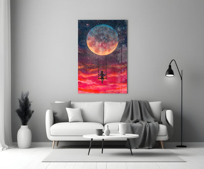 Freedom Glass Wall Art displaying a silhouetted figure on a swing beneath a large glowing moon in a vibrant red and orange sky, hung above a white sofa in a modern living room.