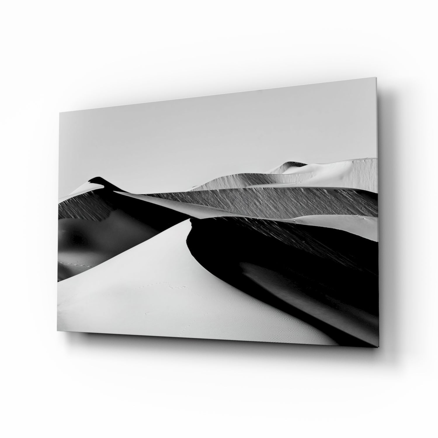 Black and white glass wall art displaying dramatic sand dunes with strong shadows and contrast, mounted on wall showing depth.