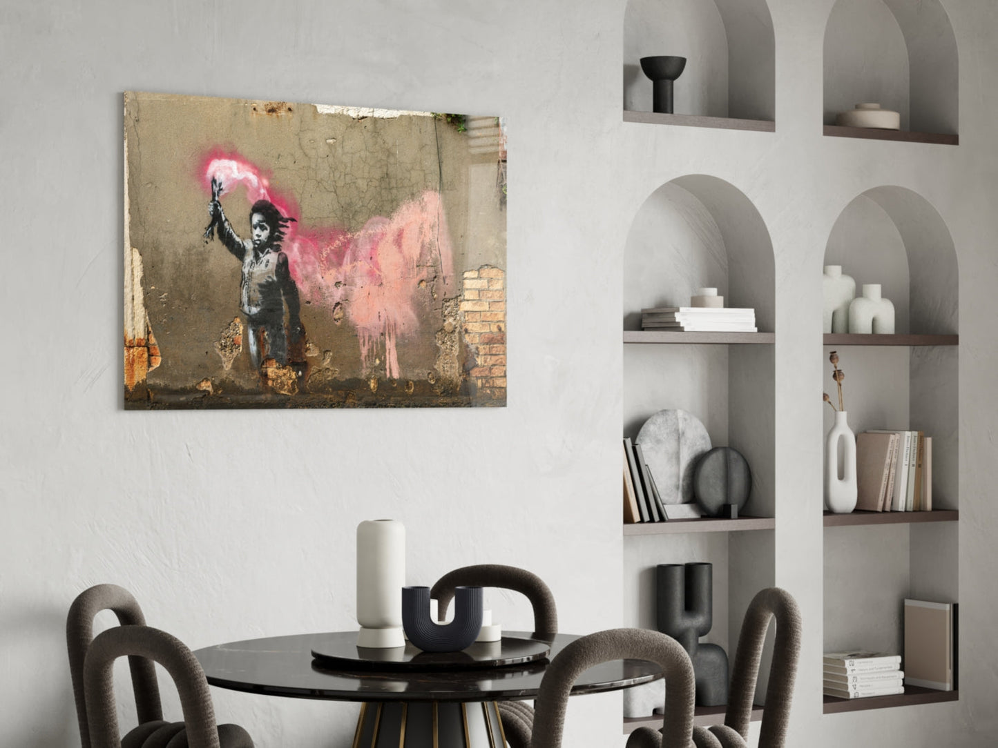 Banksy-style glass art print featuring stenciled child figure with pink spray paint, displayed above round black dining table in modern interior.