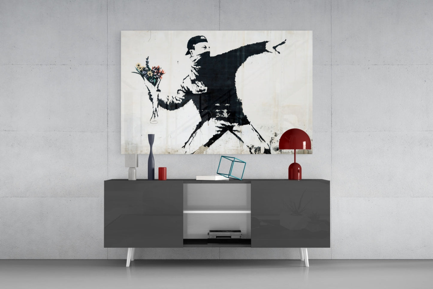 Modern living room display featuring a black and white Banksy-style stenciled artwork of a figure throwing flowers, mounted above a dark gray media console with red decorative objects.