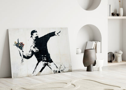 Black and white stenciled artwork of figure throwing flowers leans against white wall in minimalist interior setting.