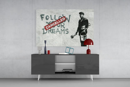 Glass art print featuring stenciled figure with "Follow Your Dreams" crossed out by red "Cancelled" banner, displayed above modern dark console table.