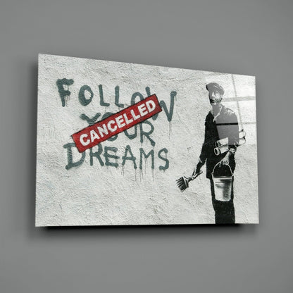 Glass art print with stenciled figure holding broom and bucket beside red "CANCELLED" stamp overlaying green text "FOLLOW YOUR DREAMS.