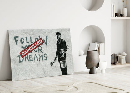 Leaning glass art displaying stenciled street art with "Follow Your Dreams" text crossed by red "Cancelled" stamp and figure holding broom, displayed in minimalist white interior.