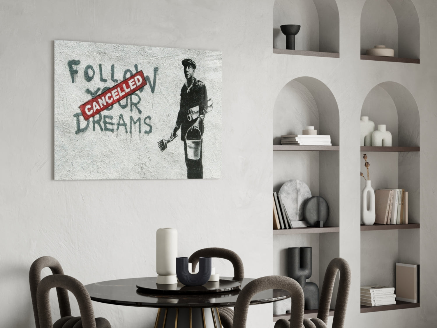 Glass art print with stenciled figure holding bucket and red "CANCELLED" banner over green text reading "FOLLOW YOUR DREAMS" displayed on white wall above dining table.