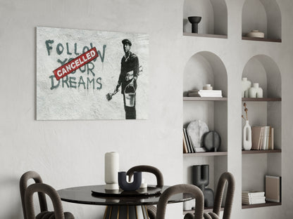 Glass art print with stenciled figure holding bucket and red "CANCELLED" banner over green text reading "FOLLOW YOUR DREAMS" displayed on white wall above dining table.