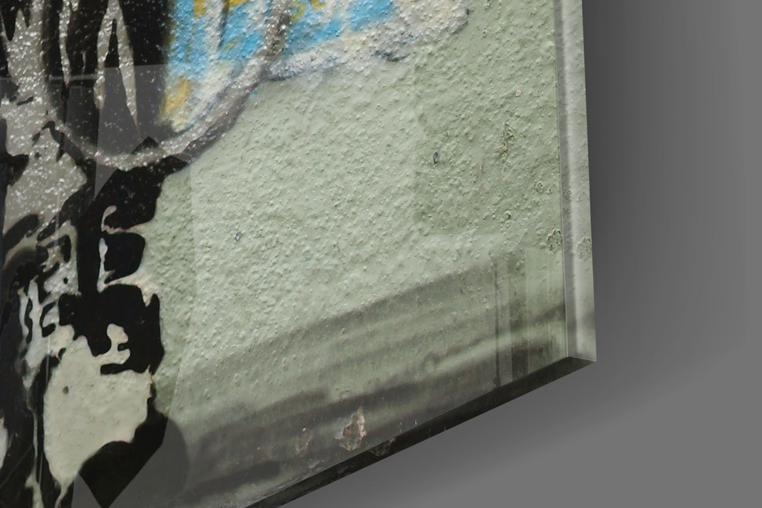 Close-up side angle of glass art panel showing textured surface with blue, yellow, and cream paint strokes and black silhouette detail.