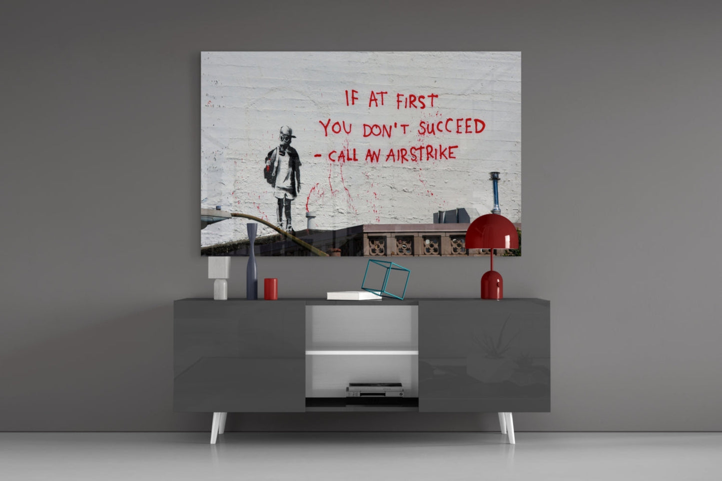 Glass art print displayed on wall above gray media console, featuring stenciled child figure and red text reading "If at first you don't succeed - call an airstrike.