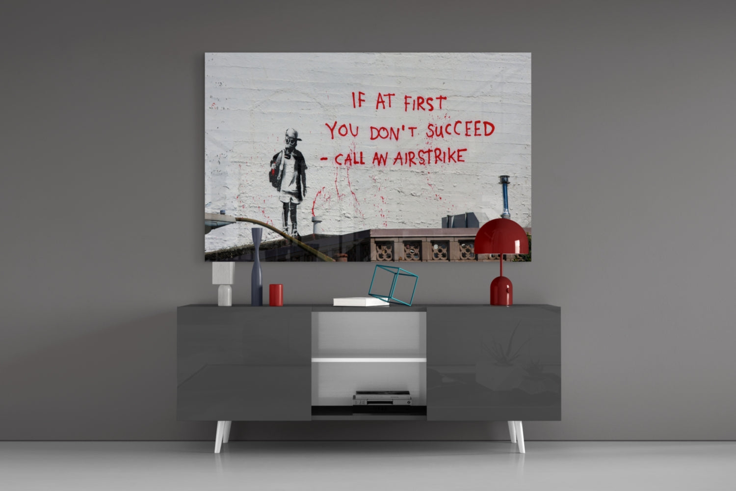Glass art print displayed on wall above gray media console, featuring stenciled child figure and red text reading "If at first you don't succeed - call an airstrike.