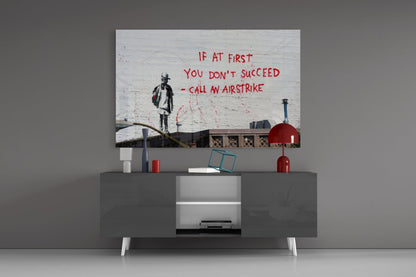 Glass art print displayed on wall above gray media console, featuring stenciled child figure and red text reading "If at first you don't succeed - call an airstrike.