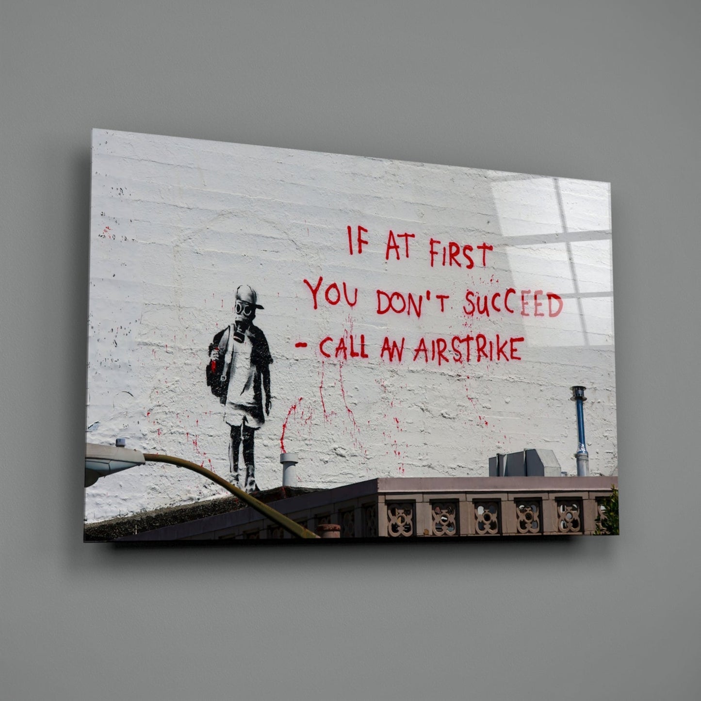 Glass art print mounted on gray wall showing street art of figure with text "IF AT FIRST YOU DON'T SUCCEED - CALL AN AIRSTRIKE" in red lettering.