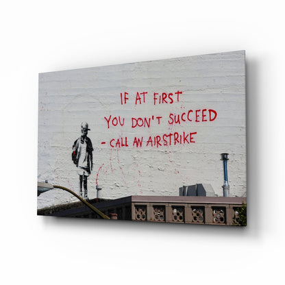Glass art canvas displaying street art featuring a stenciled figure and red text reading "If at first you don't succeed - call an airstrike" on white brick wall.