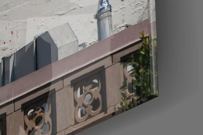 Decorative metal architectural panel with ornate circular designs mounted on brown building facade with white brick wall and metal pipe visible above.