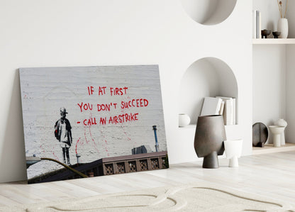 Glass art print leaning against white wall displays stenciled figure and red text "IF AT FIRST YOU DON'T SUCCEED - CALL AN AIRSTRIKE" in minimalist interior setting.
