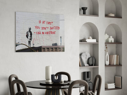 Glass art canvas displaying black silhouette figure with red text "If at first you don't succeed - call an airstrike" hanging above dining table.